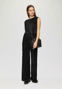 Black, sleeveless jumpsuit with a textured, vertical stripe pattern. Paired with a small black crossbody bag and chunky black shoes.
