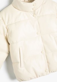Cremefarvet pufferjakke med høj krave, knaplukning og sidelommer. Fremstillet af glat, tekstureret materiale med quiltet detaljering.