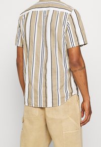 Korte mouwen, collumbia blouse met verticale strepen in beige, wit en bruin. Beschikt over een schouderstuk en een reguliere pasvorm. Katoen-linnen mix textuur.