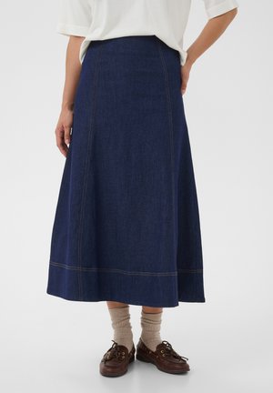 Persona con una falda larga de mezclilla azul oscuro, top blanco, calcetines beige y zapatos marrones de cuero con cordones sobre un fondo liso.