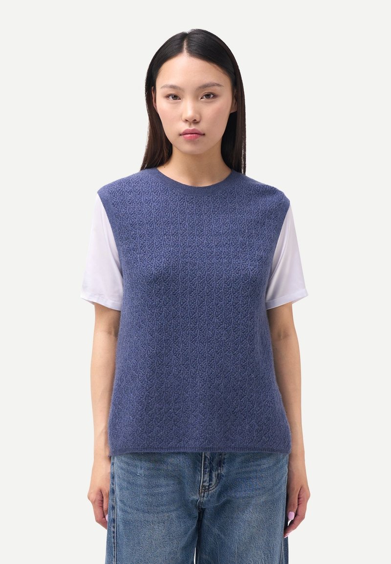 Gestrickter ärmelloser Pullover in Dunkelblau mit einer strukturierten Musterung, kombiniert mit einem weißen Hemd darunter. Legerer Schnitt mit rundem Ausschnitt.