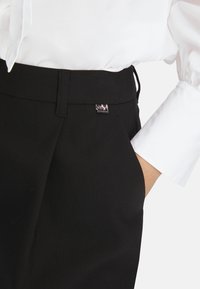 Schwarze Hose aus glattem Stoff mit flachem Bund, seitlicher Tasche und einem kleinen metallischen Markenlabel. Getragen mit einem weißen Langarmhemd.