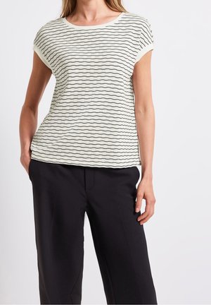 Mujer con una camiseta de punto blanca de manga corta con finas rayas negras onduladas y pantalones negros de pierna ancha, de pie con una mano en el bolsillo.