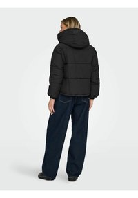 Zwarte donsjas met capuchon, voorzien van een doorgestikt ontwerp en elastische manchetten. Draagt erbij donkerblauwe jeans met wijde pijpen en zwarte laarzen.