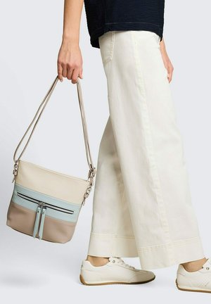 Person trägt weiße, weite Hosen und Sneakers, hält eine beige-blaue-braune Schultertasche mit Reißverschlusstaschen.