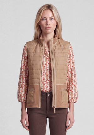 Blonde Frau trägt eine beige gesteppte Weste über einer gemusterten Bluse mit roten und weißen Formen und braunen Hosen, steht vor einem einfarbigen Hintergrund.
