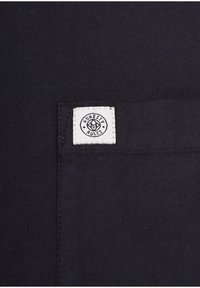 Sort stof med en firkantet hvid lap, der har teksten "ÆRLIGHED REGNER" og et cirkulært logo, syet ind i hjørnet. Flad tekstur.