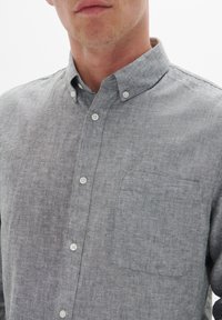 Lichtgrijs knoopshirt van een gestructureerde stof, met een gestileerde kraag en een enkele borstzak. Heeft witte knopen aan de voorkant.