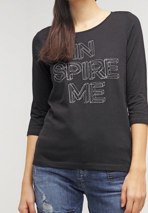 Femme portant un t-shirt à manches longues noir avec un texte en contour blanc "INSPIRE ME" et un jean bleu déchiré, debout devant un fond uni.