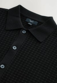 Black textured polo shirt with a ribbed collar, featuring four dark buttons and a label that reads "NEXT" in the neck area.