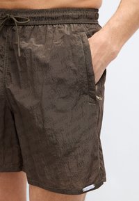 Εγγύς πλάνο ενός ατόμου που φορά καφέ σορτς Diesel με ελαστική μέση, το χέρι στην τσέπη στο πλάι με φερμουάρ, και διακριτικό λογότυπο Diesel.