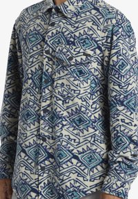 Fleece skjorte med et blåt og lilla geometrisk mønster på en cremefarvet baggrund, med knaplukning foran og en enkelt brystlomme.