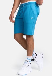 Shorts de sport turquoise avec une ceinture élastique, un cordon de serrage et des poches latérales. Détail du logo blanc sur la cuisse gauche.