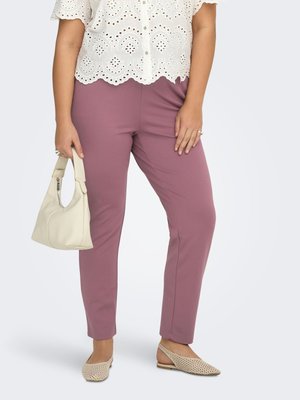 Sieviete valkā violeta pieguļošas bikses, baltu acijās iešūtu mežģīņu topu, bēša krāsas austās atvērtās kurpes ar siksniņu, rokās turot mazu bēša krāsas rokassomiņu uz baltas fona.