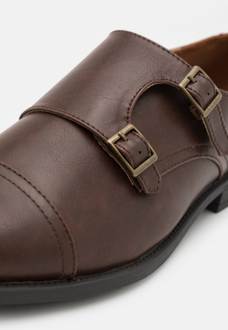 Scarpa in pelle marrone con due fibbie metalliche, una superficie liscia, punta rotonda e suola bassa in gomma nera. Dettagli cuciti sulla tomaia.