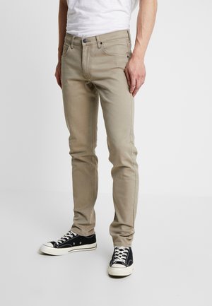 Mand iført beige slim-fit jeans, sorte og hvide sneakers og en hvid t-shirt, der står mod en ensfarvet baggrund.