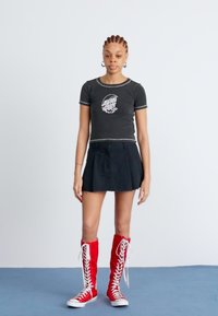 Camiseta preta curta com logo, saia preta e sapatilhas vermelhas com atacadores até ao joelho. Design simples com uma aparência casual e desportiva.
