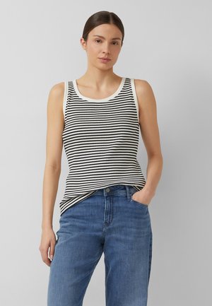 Woman wearing sleeveless black and white striped top and blue jeans, standing with one hand in pocket against plain background.