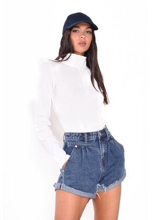 Weißes Langarmshirt mit hohem Kragen, kombiniert mit hochgeschnittenen Jeansshorts mit umgeschlagenen Säumen und Vordertaschen. Eine marineblaue Mütze rundet das Outfit ab.