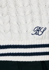 Kreemas adītas džemperis ar ribotu tumši zilu un baltu svītrojumu apakšmalā. Priekšpusē ir izšūts tumši zilais "RG" logo.