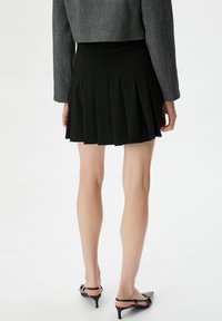Schwarzer, plissierter Minirock aus glattem Stoff, kombiniert mit einer grauen, cropped Jacke. Der Rock hat vertikale Falten für Textur und Bewegung.