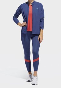 Damski strój sportowy składający się z granatowej kurtki na zamek, czerwonego topu, granatowych legginsów z czerwonymi paskami oraz białych butów sportowych.