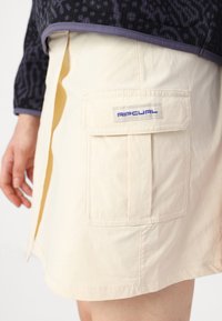 Ljusbeige kjol i mjukt tyg, med en rektangulär ficka som har en blå "Ripcurl" logotyp. Slät textur och ren fåll.