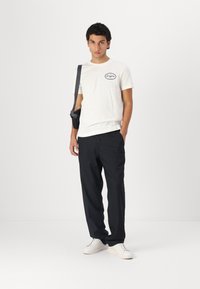 White cotton t-shirt with a black logo, paired with dark navy trousers and white sneakers. Individual stands front-facing, holding a shoulder bag.