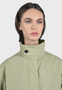 Ljusgrön, lätt jacka med hög krage, som har en svart knäppning och en minimalistisk design. Slät textur, inga synliga mönster.