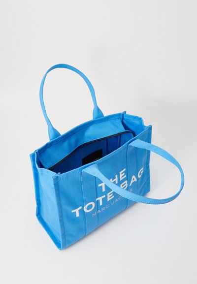 Borsa tote in tela blu con due manici, apertura superiore e grande testo bianco che recita "THE TOTE BAG MARC JACOBS." Interno visibile.