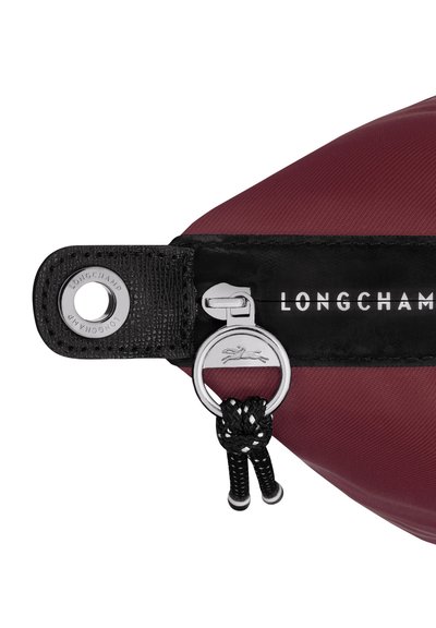 Dunkelroter Nylonbeutel mit schwarzem, strukturiertem Besatz, ausgestattet mit silbernem Reißverschluss und auffälligem Logo. Runde Metallösen-Akzentuierung inklusive.