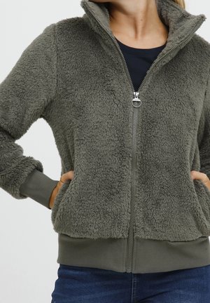 Frau trägt eine grau-grüne, flauschige Jacke mit Reißverschluss und die Hände in den Taschen, kombiniert mit dunkelblauen Jeans und einem schwarzen Shirt darunter.