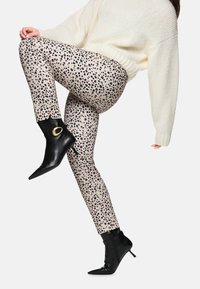 Leopardenmuster Leggings in einem hellbeigen Grundton mit schwarzen Flecken, kombiniert mit schwarzen spitzen Stiefeletten, die ein goldfarbenes Akzentdetail aufweisen.
