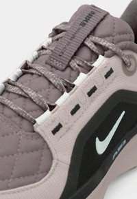 Nike superge v mehki roza barvi z nabranim vzorcem, s črnimi poudarki, belimi vezalkami in izrazitim Swoosh logotipom ob strani.