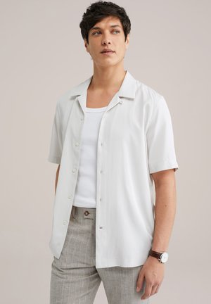 Chemise blanche à manches courtes, boutonnée, en tissu lisse avec une coupe décontractée. Elle présente un col et une légère brillance, associée à un pantalon gris clair texturé.