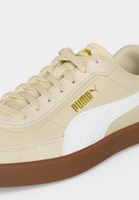 Μπεζ αθλητικό παπούτσι Puma με λευκή πλευρική ρίγα, χρυσό λογότυπο Puma, μπεζ κορδόνια και καφέ ελαστική σόλα, εμφανιζόμενο από πάνω πλάγια γωνία.