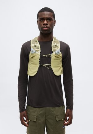 Young man wearing a black long-sleeve shirt and olive cargo shorts with a beige hydration vest holding two water bottles.