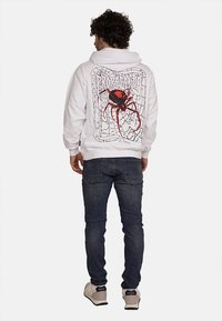 Felpa bianca con cappuccio caratterizzata da un design grafico di un ragno nero con dettagli rossi, stampato sulla schiena su uno sfondo di ragnatela, in materiale di cotone.
