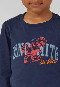 Langarmshirt in Marineblau mit einem roten Dinosaurier-Grafik, der einen Fußball tritt, und hellblauem sowie orangefarbenem Text, der "DINOMITE Dribbler" lautet.