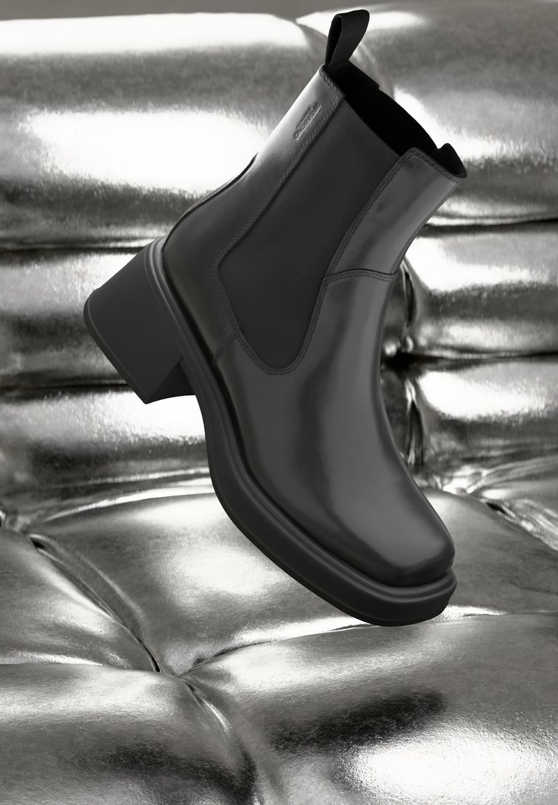 Bottine noire en cuir avec talon épais et panneaux latéraux élastiques reposant sur une surface argentée matelassée et réfléchissante.