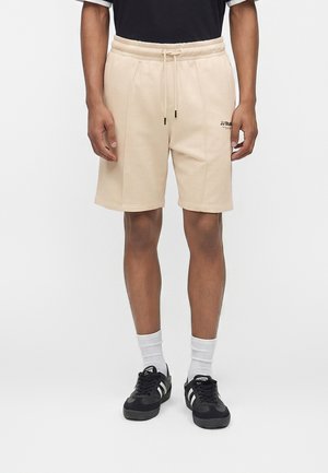 Férfi visel bézs, húzózsinóros rövidnadrágot a JJ Studio logóval, fekete cipő fehér csíkkal, fehér zoknival, egyszínű háttér előtt áll.