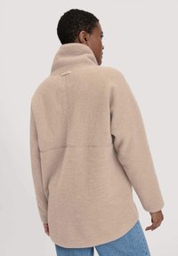 Giacca in fleece beige con colletto alto, maniche lunghe e taglio dritto. Presenta un sottile dettaglio di cucitura e un'etichetta sul retro.