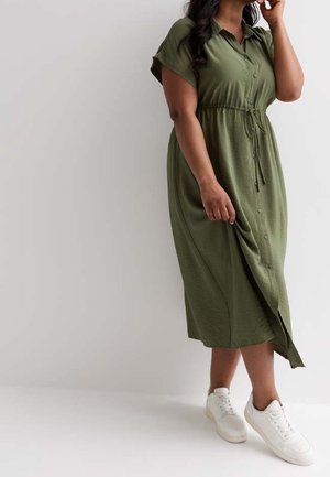 Kvinde iført en olivengrøn midi-kjole med knapper foran, taljebånd og hvide sneakers, stående mod en ensfarvet lys baggrund.