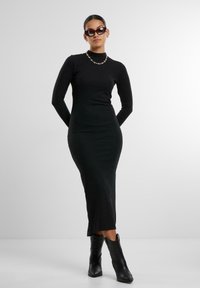 Vestito nero a maniche lunghe, a coste, con collo alto, silhouette aderente e uno spacco laterale, abbinato a stivaletti neri alla caviglia e occhiali da sole.
