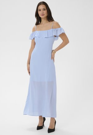 Femme portant une robe maxi bleu clair à épaules dénudées avec des volants, des talons noirs et un bracelet en or, debout devant un fond uni.