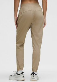 Beige sportbroek met een gladde textuur, elastische tailleband en ritssluitingen bij de enkel, gecombineerd met wit-zwart sneakers.