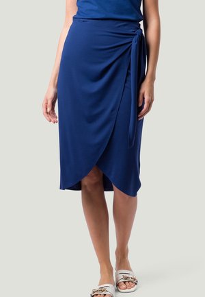 Femme portant une jupe portefeuille bleu marine longueur genou avec un lien sur le côté et des sandales blanches à enfiler avec un détail chaîne dorée.
