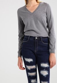 Femme portant un pull long à col en V gris et un jean taille haute bleu foncé usé avec plusieurs déchirures et des patches effilochés.