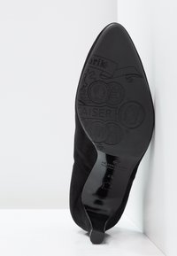 Chaussure à talon haut noire en suède avec un bout pointu et une silhouette élégante. Semelle détaillée avec motifs en relief et marque. Fini lisse et mat.