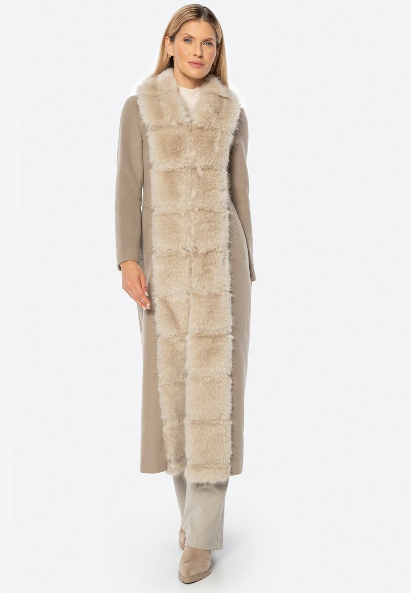 Lange beige jas met nep bontkraag en voorpaneel. Textuur van het bont contrasteert met de gladde stof. Model poseert in bijpassende lichte broek en schoenen.